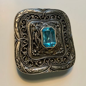 May include: A silver-toned brooch with an ornate design and a light blue gemstone in the center.