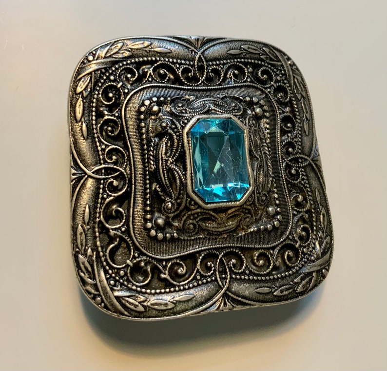May include: A silver-toned brooch with an ornate design and a light blue gemstone in the center.