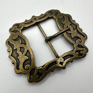 May include: A gold-toned metal belt buckle with an ornate, intricate design. The buckle has a rectangular shape with a detailed, floral pattern.