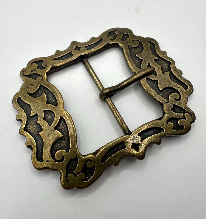May include: A gold-toned metal belt buckle with an ornate, intricate design. The buckle has a rectangular shape with a detailed, floral pattern.