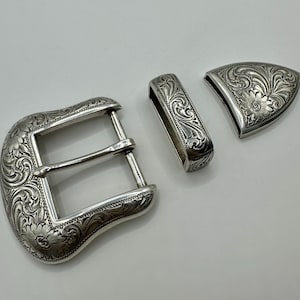 Op de afbeelding: Een zilverkleurige gesp set met ingewikkelde bloemen- en scrollgravure. De set bevat een gesp, een riemlus en een decoratief eindstuk, allemaal met een bijpassend ontwerp. De stukken zijn waarschijnlijk van metaal.