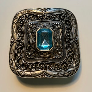 May include: A silver-toned square-shaped brooch with an ornate design and a light blue gemstone in the center.
