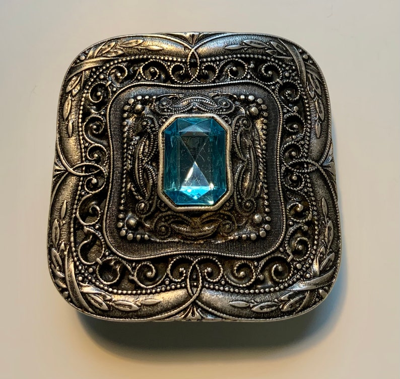 May include: A silver-toned square-shaped brooch with an ornate design and a light blue gemstone in the center.