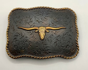 May include: A gold and black belt buckle with a detailed floral design and a gold longhorn steer head in the center.