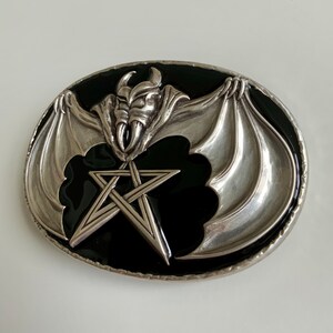 May include: Silver belt buckle with a black enamel background. The buckle features a silver bat with wings spread wide, holding a five-pointed star in its claws.