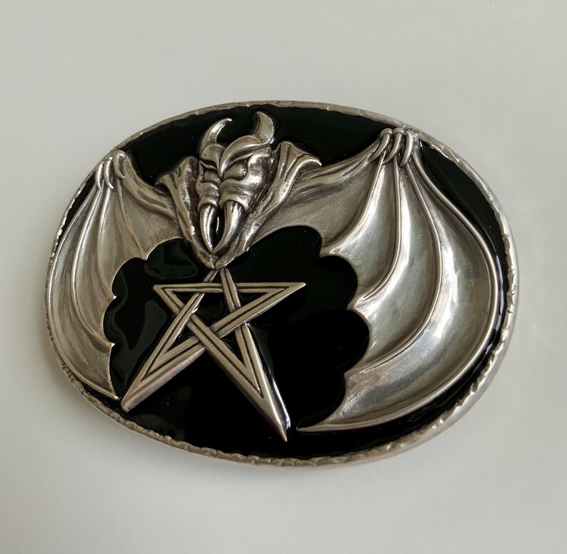 May include: Silver belt buckle with a black enamel background. The buckle features a silver bat with wings spread wide, holding a five-pointed star in its claws.