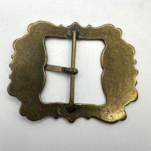 May include: A gold-toned metal belt buckle with a decorative, ornate design. The buckle has a rectangular shape with a smooth, polished finish.