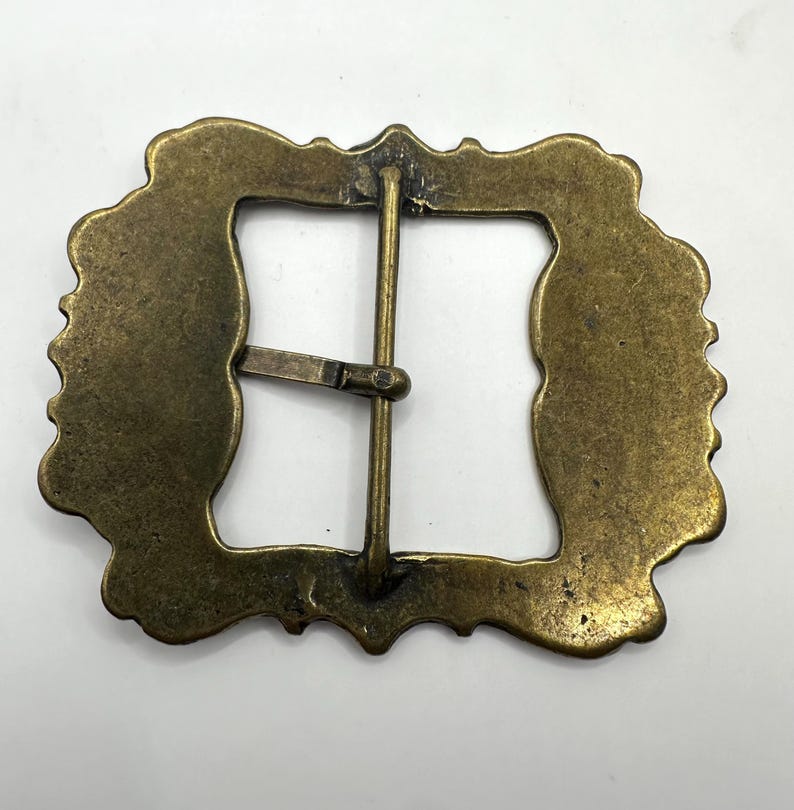 May include: A gold-toned metal belt buckle with a decorative, ornate design. The buckle has a rectangular shape with a smooth, polished finish.