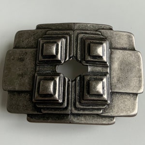 May include: A silver-toned metal belt buckle with a geometric design. The buckle has a square shape with four smaller squares in the center, each with a raised, textured surface.