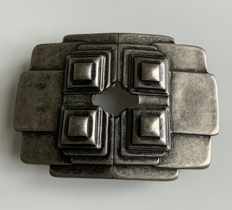 May include: A silver-toned metal belt buckle with a geometric design. The buckle has a square shape with four smaller squares in the center, each with a raised, textured surface.