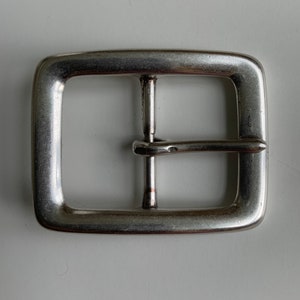 May include: A rectangular silver-toned metal belt buckle with rounded corners. The buckle features a central vertical bar and a hinged prong that extends across the frame, designed to secure a belt strap through the open rectangular space.