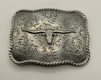 May include: Silver rectangular belt buckle with a longhorn steer head. The buckle has detailed floral and scroll patterns, and a rope-style edge. A Western-style accessory, suitable for a belt.