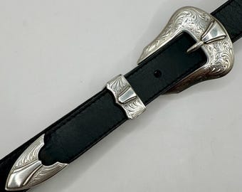May include: A black leather belt with a silver buckle featuring an intricate floral design. The buckle is engraved with a detailed pattern.