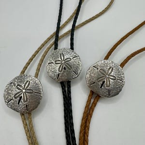 Op de afbeelding: Drie zilverkleurige bolo-stropdassen met zeesterontwerpen. Elke stropdas heeft een ronde, getextureerde hanger met een gedetailleerd bloemenpatroon. De koorden zijn gevlochten in beige, zwart en bruin.