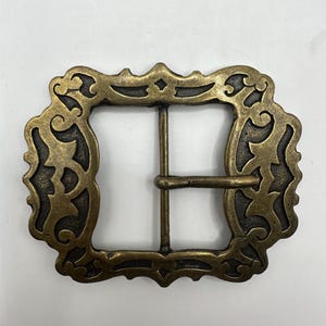 May include: A gold-toned metal belt buckle with an ornate, intricate design. The buckle has a square shape with a decorative border and a center opening for a belt.