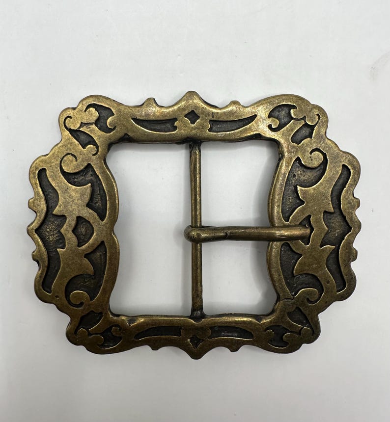 May include: A gold-toned metal belt buckle with an ornate, intricate design. The buckle has a square shape with a decorative border and a center opening for a belt.