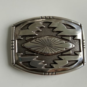 Könnte beinhalten: Eine silberne Gürtelschnalle mit geometrischem Design. Die Schnalle hat eine zentrale ovale Form mit einem Sonnenstrahlenmuster und ist von einem sich wiederholenden Muster aus Dreiecken umgeben.