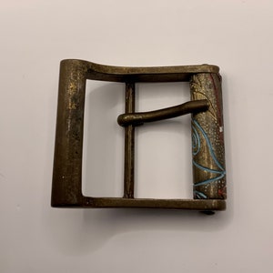 May include: A brass belt buckle with a decorative, colorful design. The buckle has a rectangular shape with a curved top and a metal loop on the side.