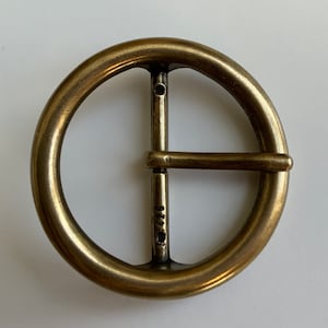 May include: A brass belt buckle with a round, smooth, polished surface and a simple, straight, metal bar closure. The buckle has the number "2416" stamped on the bar.