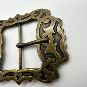 May include: A gold-toned metal belt buckle with an ornate, intricate design. The buckle has a rectangular shape with a detailed, scroll-like pattern.