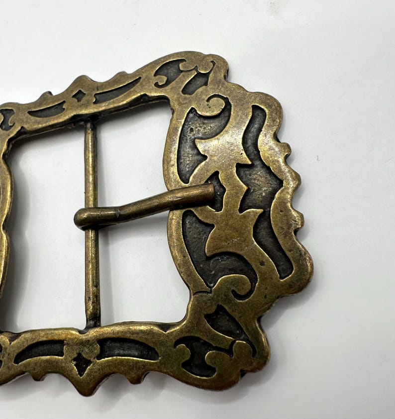 May include: A gold-toned metal belt buckle with an ornate, intricate design. The buckle has a rectangular shape with a detailed, scroll-like pattern.