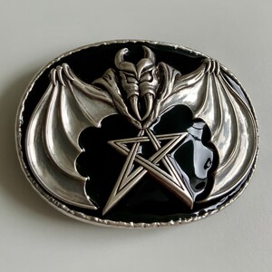 May include: Silver belt buckle with a black enamel background. The buckle features a pentagram with a bat-like creature with wings and horns in the center.