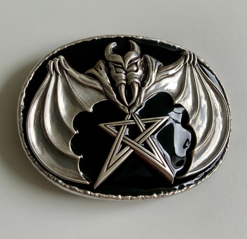 May include: Silver belt buckle with a black enamel background. The buckle features a pentagram with a bat-like creature with wings and horns in the center.