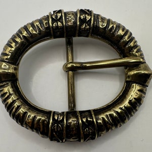 May include: A gold-toned oval belt buckle with a textured, ornate design. The buckle has a detailed, raised pattern of lines and circles.