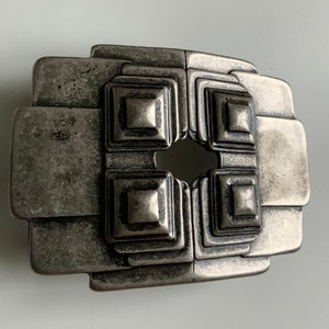 May include: A silver-toned metal belt buckle with a geometric design. The buckle has a square shape with a central opening and a series of concentric squares around the opening.