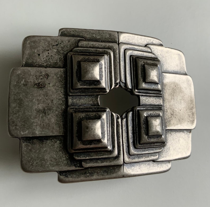 May include: A silver-toned metal belt buckle with a geometric design. The buckle has a square shape with a central opening and a series of concentric squares around the opening.