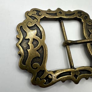 May include: A gold-toned metal belt buckle with an ornate, intricate design. The buckle has a rectangular shape with a detailed, decorative border.