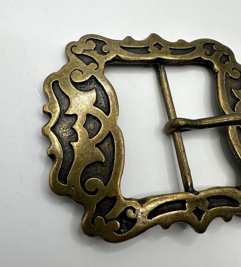 May include: A gold-toned metal belt buckle with an ornate, intricate design. The buckle has a rectangular shape with a detailed, decorative border.