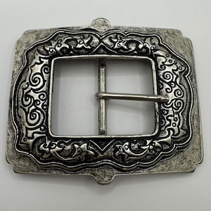 May include: A silver-toned belt buckle with an ornate design featuring intricate swirls and floral patterns.