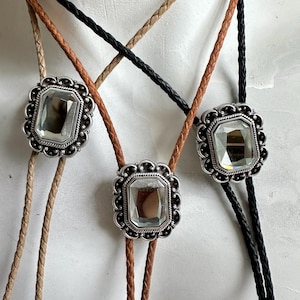 Op de afbeelding: Drie bolo-strikken met verschillende gekleurde koorden. Elke stropdas heeft een zilverkleurige, sierlijke, rechthoekige hanger met een heldere, gefacetteerde edelsteen.