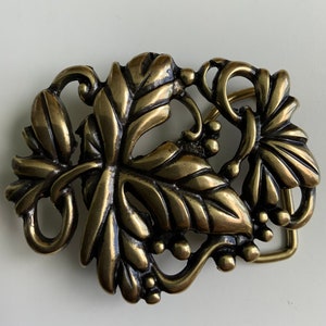 Puede incluir: Una hebilla de cinturón de metal dorado con un diseño detallado de hojas y vides.