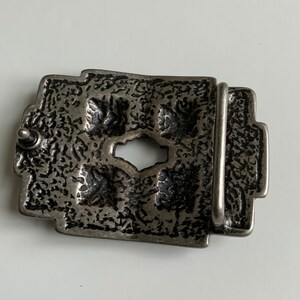 May include: A silver-toned belt buckle with an intricate design. The buckle has a textured surface and a rectangular shape.