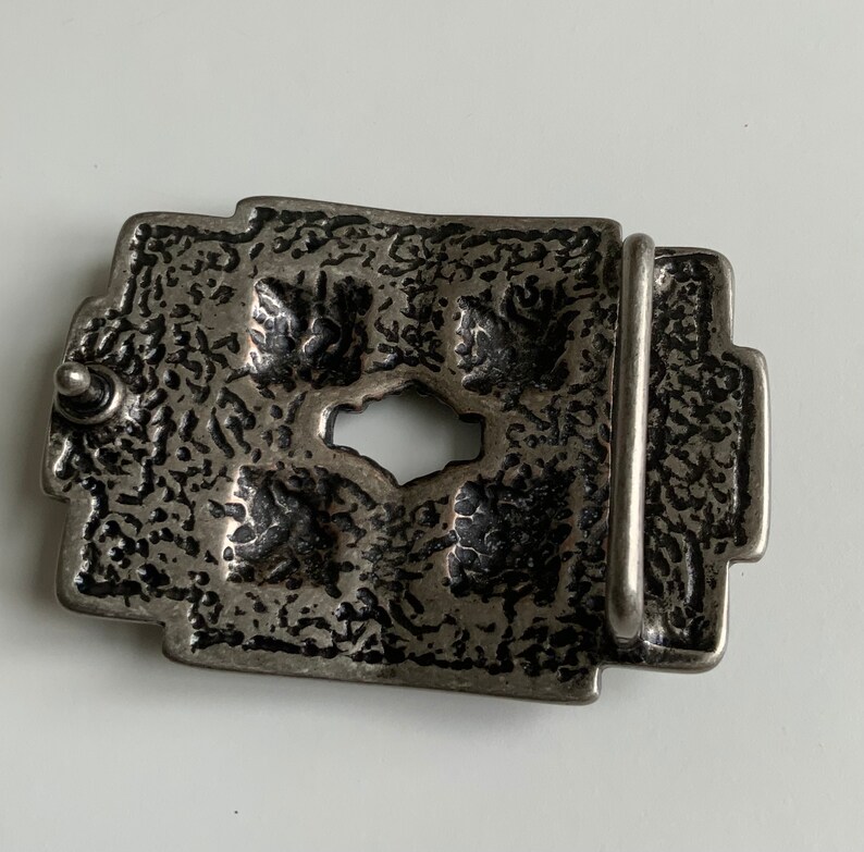 May include: A silver-toned belt buckle with an intricate design. The buckle has a textured surface and a rectangular shape.