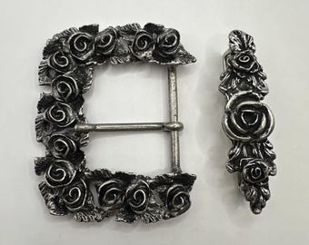 May include: A silver-toned metal belt buckle and matching keeper. The buckle is rectangular, adorned with detailed rose and leaf designs around the frame. The keeper has a single rose at the center.