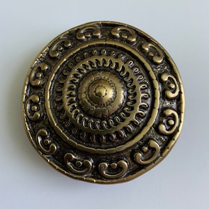 May include: A round, gold-toned metal decorative piece with intricate circular patterns. The centre of the piece features a raised, circular design with a textured surface.