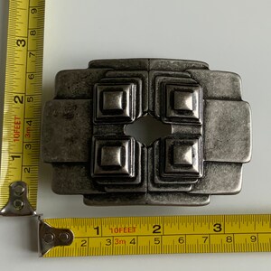 May include: A silver-toned metal belt buckle with a geometric design. The buckle has a square shape with four smaller squares inside, each with a raised center. The buckle has a distressed finish, giving it a vintage look.