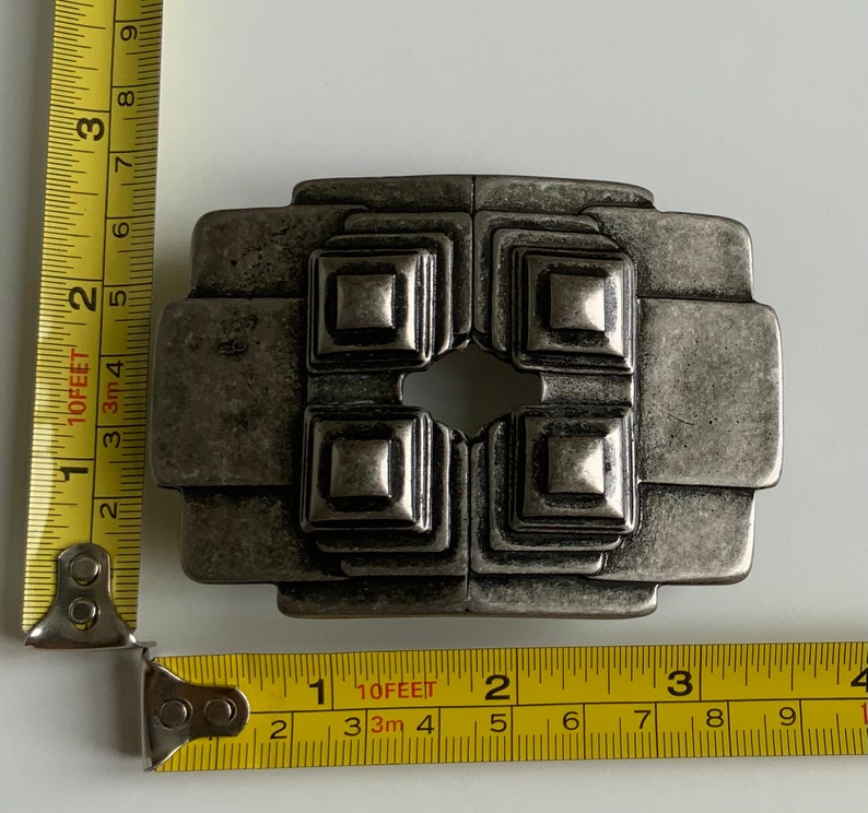 May include: A silver-toned metal belt buckle with a geometric design. The buckle has a square shape with four smaller squares inside, each with a raised center. The buckle has a distressed finish, giving it a vintage look.