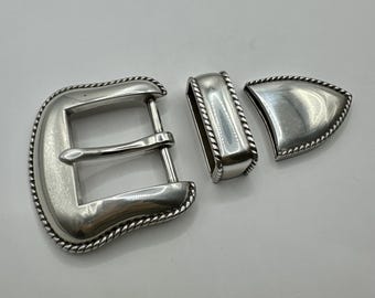 May include: Silver metal belt buckle set. The set includes a rectangular buckle, a belt loop, and a decorative piece. Each piece has a rope-like border. The buckle is a classic western style.