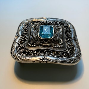May include: A silver belt buckle with an ornate design and a blue gemstone in the center.