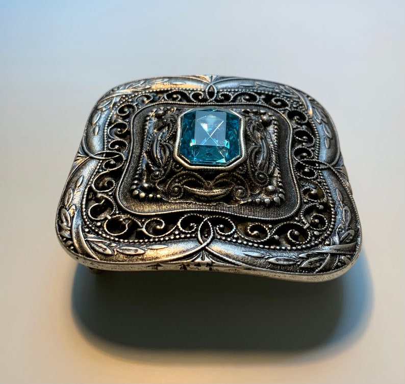 May include: A silver belt buckle with an ornate design and a blue gemstone in the center.