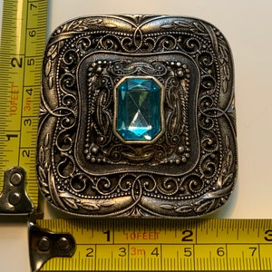 May include: A silver-toned brooch with an ornate, filigree design. The brooch features a large, light blue gemstone in the center.