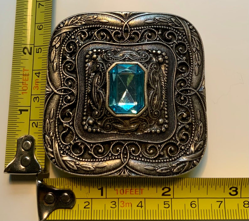 May include: A silver-toned brooch with an ornate, filigree design. The brooch features a large, light blue gemstone in the center.