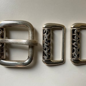 May include: Three silver-toned metal buckles with decorative floral filigree. The largest buckle is square with a central bar, while the other two are rectangular. The buckles are likely for belts or other accessories.