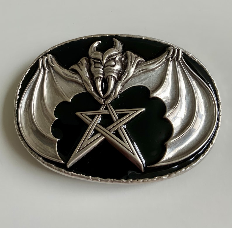 May include: Silver belt buckle with a black enamel background. The buckle features a silver bat with wings spread wide and a pentagram in the center.