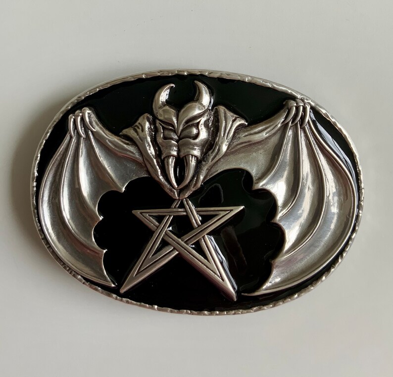 May include: Silver belt buckle with a black enamel background. The buckle features a bat with wings spread wide and a pentagram in the center.