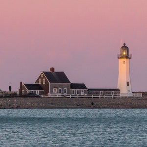 Op de afbeelding: Een witte vuurtoren met een zwart dak en een licht bovenop, die op een rotsachtige kust staat met een klein huis op de voorgrond. De lucht is zacht roze van kleur.